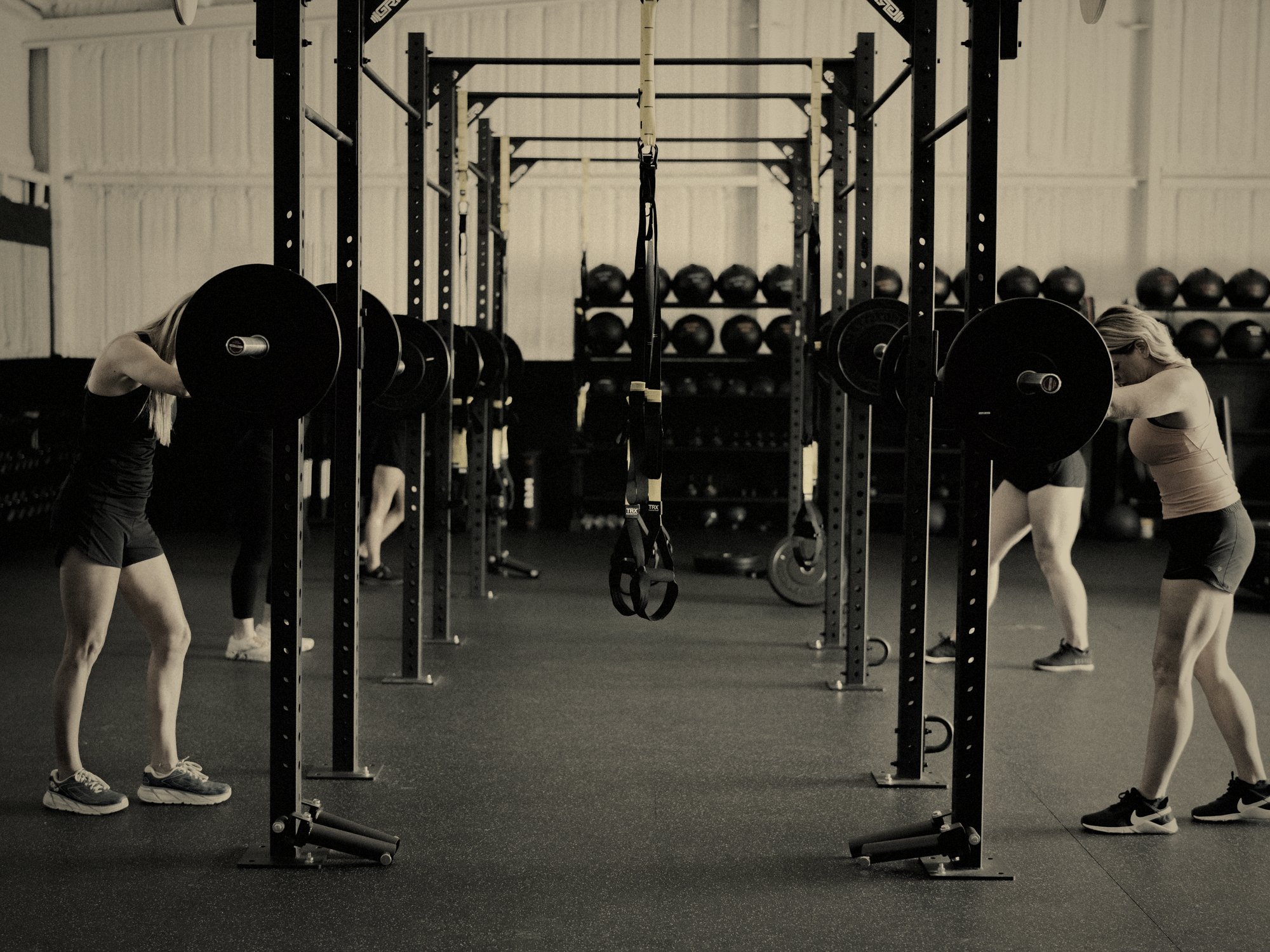 Members stretching against loaded barbells between rows of squat racks at M6 Fit